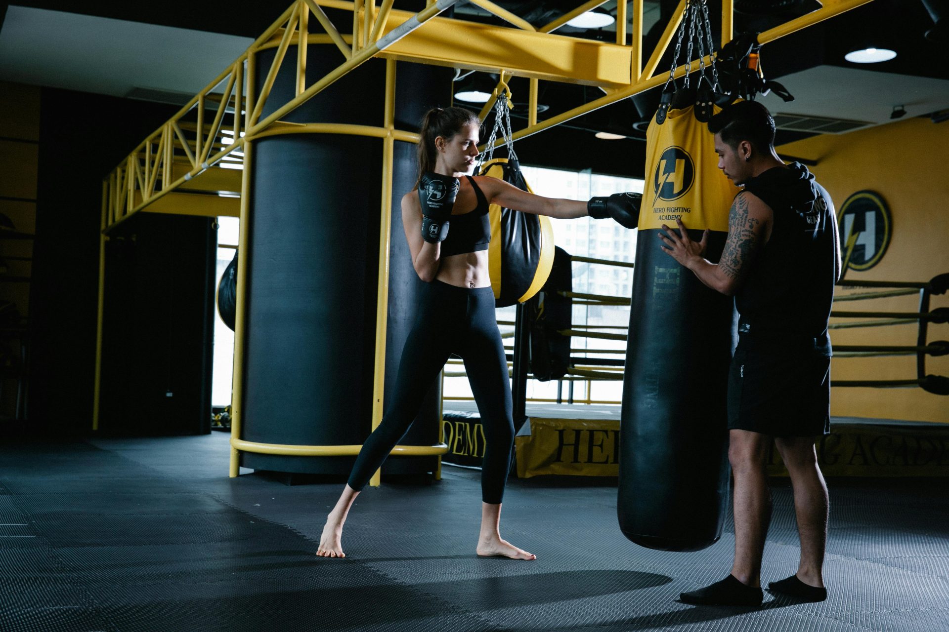 Cuerpo completo de una luchadora deportiva golpeando un saco de boxeo durante un entrenamiento con un instructor latinoamericano personal tatuado en un gimnasio moderno